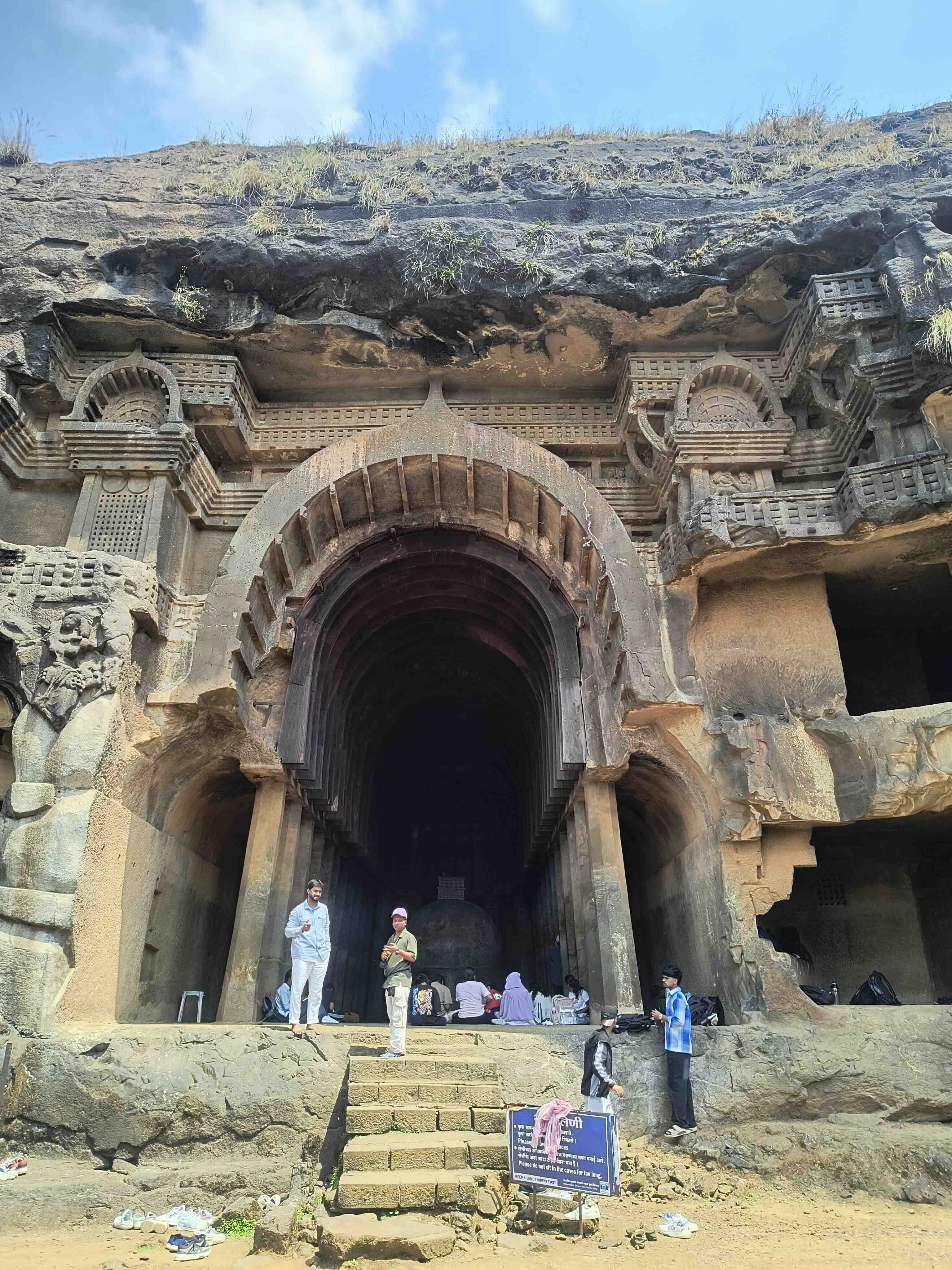 The arched main chaitya entrance of the Bhaje caves, cut into a dark rock face, with visitors standing on the steps and seated inside.