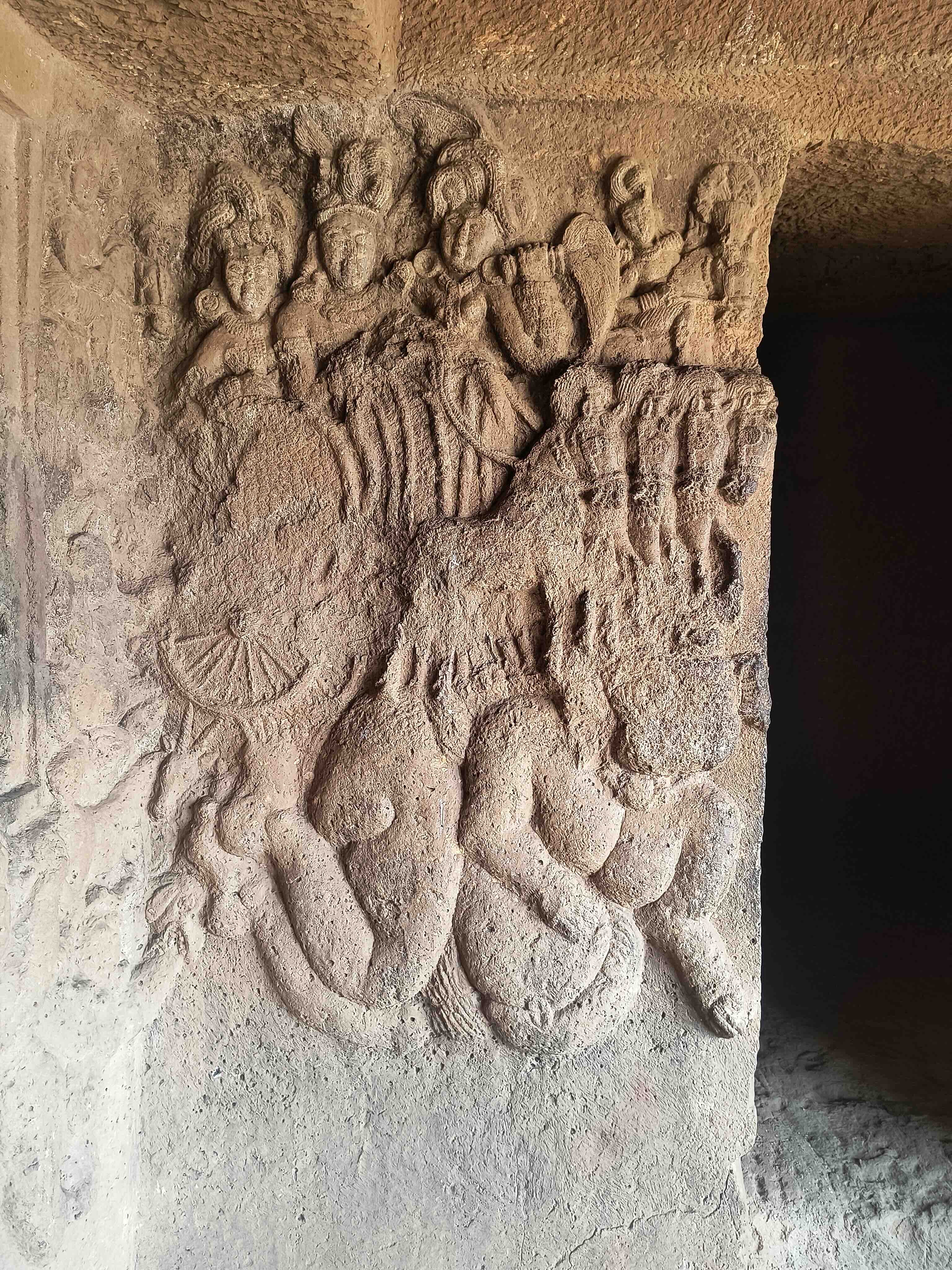 A weathered relief in a Bhaje cave showing the sun god's chariot drawn by horses, with attendant figures above and a dark doorway at right.
