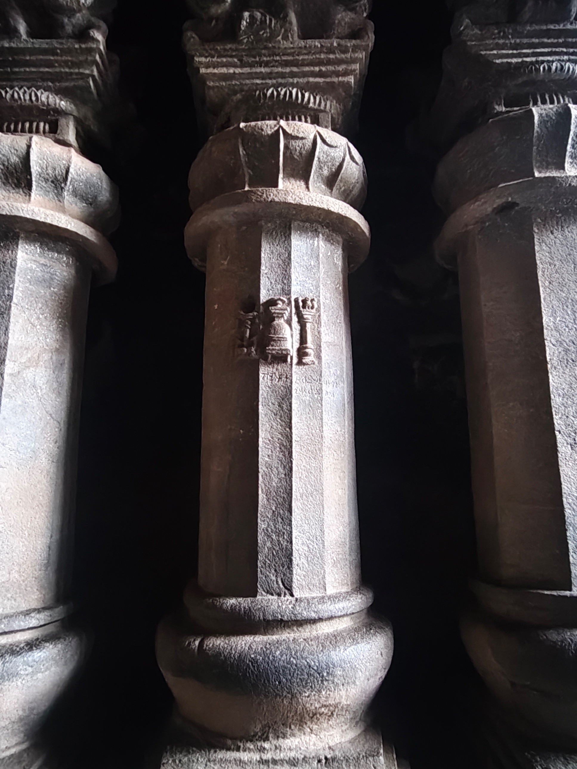 A carved pillar inside the Karle hall with a pot-on-base motif flanked by two small pillar motifs, an inscription beneath, and a lotus-carved capital above.