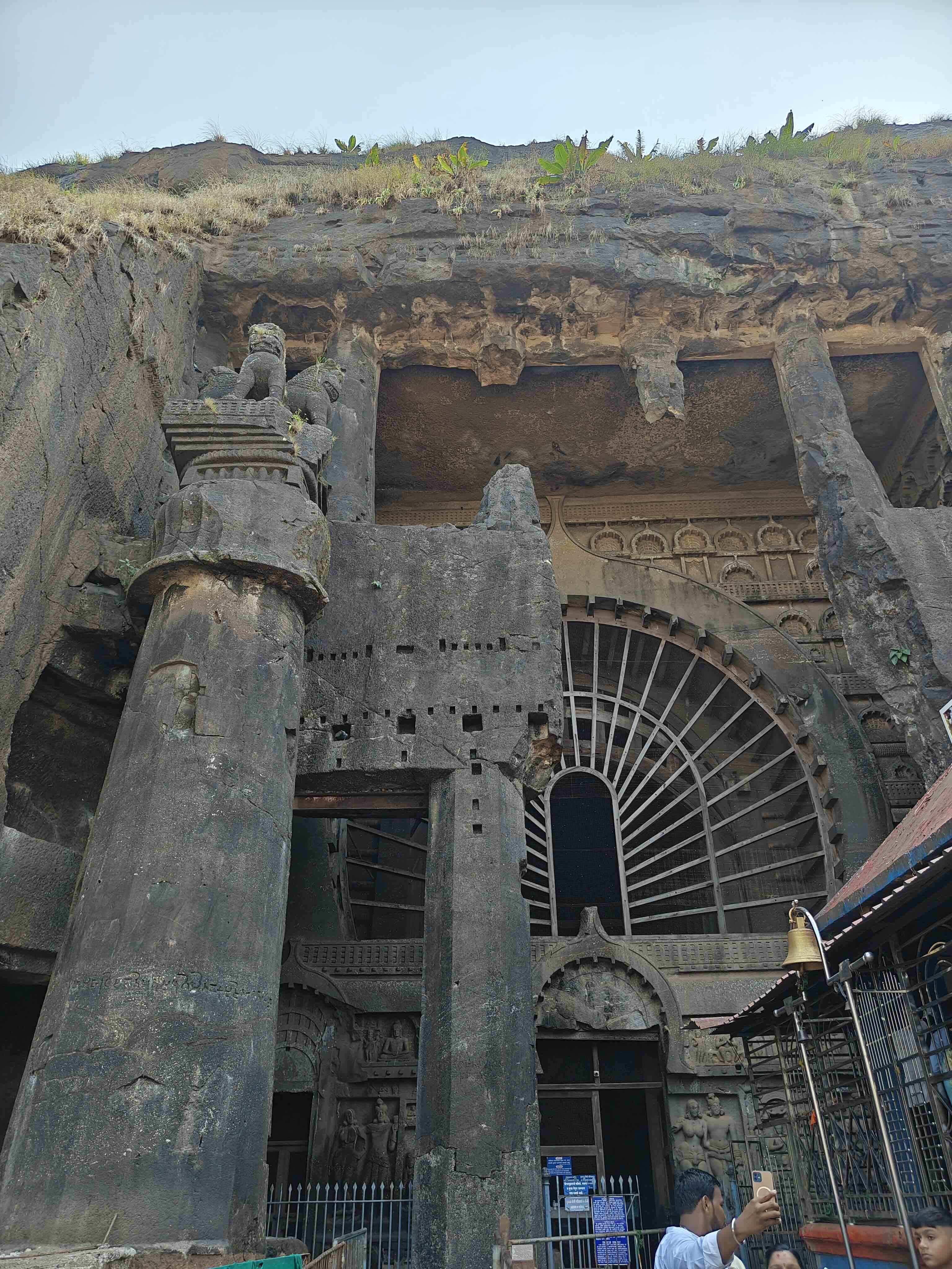 A tall stone pillar topped with a lion capital stands before the arched, sunburst-windowed facade of the Karle Chaitya Griha, with a temple bell at right.